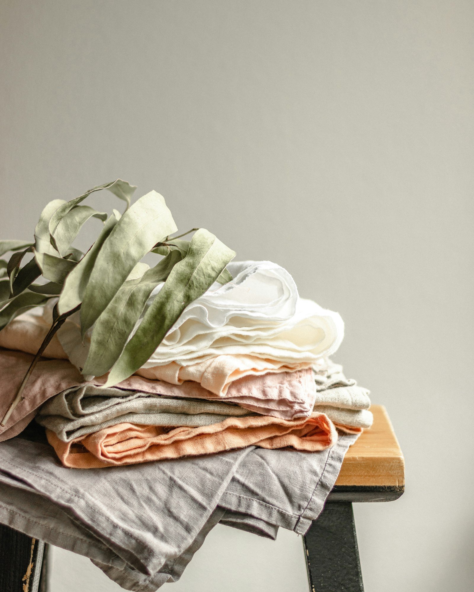 Du linge de lin plié en pile sur un tabouret en bois, sous une branche de feuillage séché